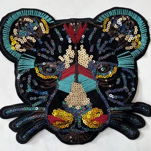 Puede incluir: Una aplicación de fieltro negro de un rostro de tigre con lentejuelas y cuentas de colores. Los ojos del tigre están delineados en turquesa y la nariz es roja.