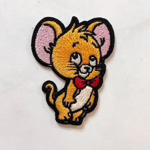 May include: Embroidered patch featuring a cartoon mouse with yellow fur, pink ears, and a red bow tie. The mouse has a white belly and is outlined in black. This patch is suitable for clothing or accessories.