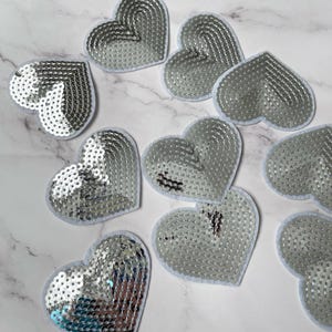 May include: Ten silver sequin heart patches with white felt backing. The hearts are various sizes and are arranged in a random pattern.