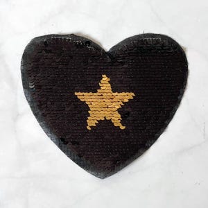 May include: A heart-shaped patch with black sequins and a gold star. The patch is designed for decorative use, possibly to embellish clothing or accessories. The sequins provide a shimmering, eye-catching effect.