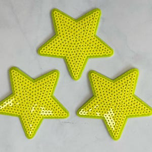 May include: Three yellow star-shaped patches made of sequins. The stars are on a white background.