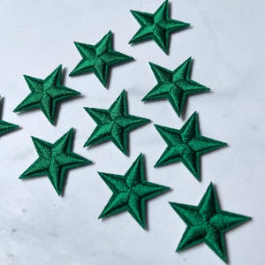 Puede incluir: Un conjunto de parches bordados en forma de estrella de color verde esmeralda. Cada estrella tiene cinco puntas y está rellena con costuras detalladas. Estos parches son ideales para añadir un toque decorativo a ropa, bolsos u otros artículos de tela.