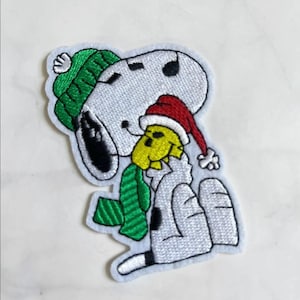 May include: Embroidered patch featuring Snoopy in a winter outfit, wearing a green knit cap and scarf, and a red Santa hat. The patch has a white background and is suitable for clothing or accessories.