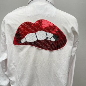 May include: White long-sleeved button-down shirt with a red sequined lips design on the back.