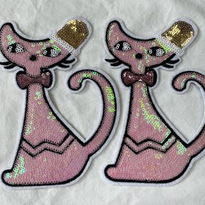 May include: Two pink sequin cat patches with black outlines. Each cat wears a gold sequin hat with white trim and a maroon bow tie. The cats have large eyes and a curled tail.
