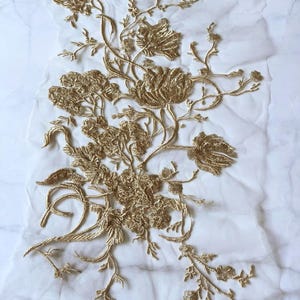 Peut inclure: Applique de broderie florale dorée sur un fond de tulle blanc. Le motif présente des fleurs, des feuilles et des tiges détaillées, créant un motif délicat et orné. Idéal pour les vêtements ou les projets d'artisanat.