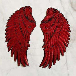 May include: Two red sequined wing patches with black outlines. The wings are detailed with individual feather shapes and are symmetrical. The patches are likely for decorative purposes, such as embellishing clothing or accessories.