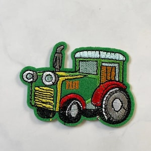May include: Embroidered green tractor patch with yellow, red, and black accents. The patch features a detailed design of a tractor with a smokestack, wheels, and headlights. This patch is suitable for clothing or accessories.