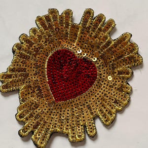 May include: A heart-shaped patch with a red center and a gold sequined border. The gold sequins are arranged in a sunburst pattern, and the patch has a black backing. This decorative patch can be used for clothing or accessories.