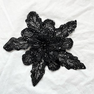 May include: A black floral applique with intricate sequin and bead detailing. The applique features multiple petals radiating from a central point, creating a flower-like design. The sequins add a shimmering effect to the fabric.