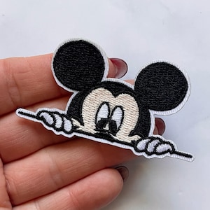 May include: Embroidered iron-on patch featuring a cartoon Mickey Mouse peeking over a white banner. Mickey Mouse has black ears and a beige face with black eyes.