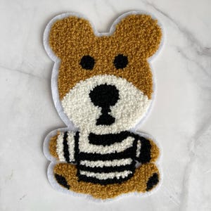 May include: A brown teddy bear with a white and black striped shirt. The bear is made of a fluffy material and has a white face with black eyes and a black nose.