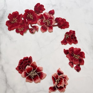 May include: A collection of embroidered floral appliques in red and pink hues, featuring gold centres. The appliques vary in size and arrangement, ideal for craft projects or garment decoration. The detailed stitching provides a textured, three-dimensional appearance.