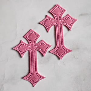 Może przedstawiać: Dwie różowe naszywki w kształcie krzyża z dekoracyjnym wzorem. Każda naszywka ma białą, szytą obwódkę. Krzyże wykonane są z miękkiego materiału i prezentowane na białej powierzchni.