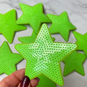 Puede incluir: Un grupo de parches en forma de estrella verde lima hechos de lentejuelas. Los parches están dispuestos en semicírculo con la estrella más grande en el centro.