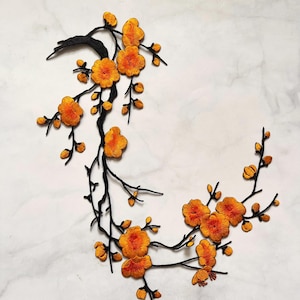Puede incluir: Rama floral bordada con flores y capullos naranjas, sobre un fondo de mármol blanco. Las ramas negras se curvan con gracia, creando un diseño decorativo. Las flores tienen costuras detalladas.