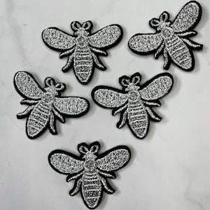 May include: Five silver glitter bee iron-on patches. The bees are outlined in black and have a detailed design.