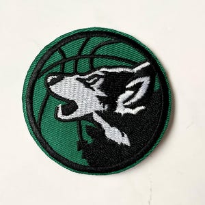 Könnte beinhalten: Grün-schwarzer gestickter Aufnäher mit einem heulenden Wolf in einem Basketballkorb.