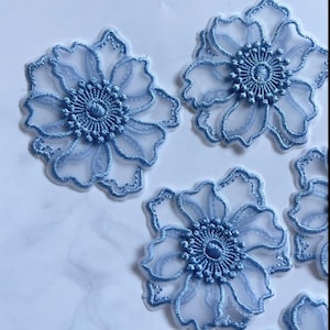 Puede incluir: Varias aplicaciones florales en azul claro. Cada aplicación presenta un contorno bordado detallado y un diseño central texturizado. Los pétalos son translúcidos, creando un efecto delicado y en capas. Adecuado para diversos proyectos de manualidades.