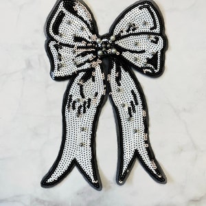 Può includere: Un fiocco bianco di paillettes con contorno nero e un centro di perla. Il fiocco è un'applicazione in tessuto.