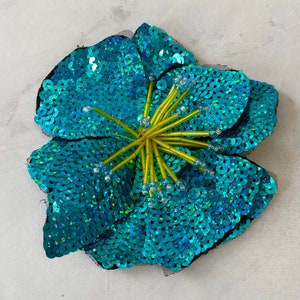 May include: A turquoise blue flower made of sequins with a yellow center. The flower is a decorative embellishment.