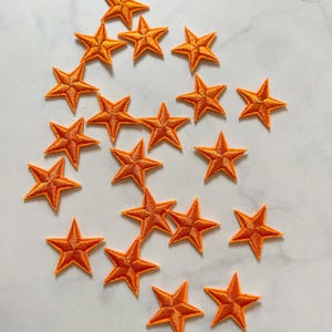 May include: A collection of orange star-shaped embroidered patches. Each star has five points and a detailed stitched outline. The patches are scattered on a light-colored surface, suitable for embellishing clothing or accessories.