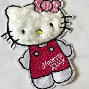 Peut inclure: Écusson brodé blanc et rose représentant un chat de dessin animé avec un nœud rose. Le chat porte une robe rose avec le texte "Heeey Kitty". Contour noir.