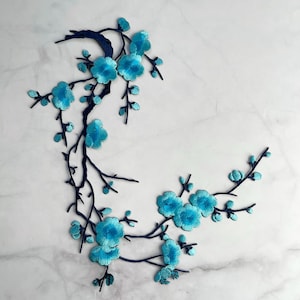 Könnte beinhalten: Gestickte Blumenapplikation mit einem Zweig mit türkisfarbenen Blüten und Knospen. Die Blüten haben ein strukturiertes Aussehen, und die Zweige sind dunkelmarineblau. Die Applikation ist vor einem weißen Marmorhintergrund angeordnet.