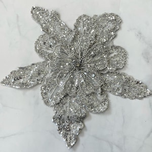 Puede incluir: Una aplicación de flor de lentejuelas plateadas con un diseño delicado e intrincado. La flor está hecha de pequeñas lentejuelas brillantes que crean un efecto brillante.