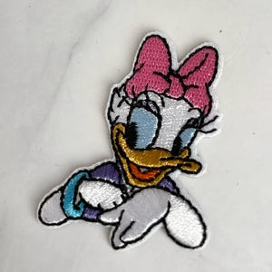 Könnte beinhalten: Ein gestickter Bügelaufkleber mit einem Cartoon-Bild eines weißen Entchens mit einer rosa Schleife, blauen Augen und einem gelben Schnabel. Das Entchen trägt ein lila-blaues Hemd und hat ein türkisfarbenes Armband am linken Flügel.