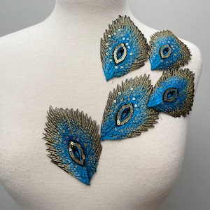 May include: Five peacock feather-shaped appliques with gold and blue embroidery. The appliques are made of fabric and have a shiny, iridescent finish.