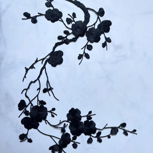 Könnte beinhalten: Schwarze florale Stickerei Applikation mit einem Zweig mit Blumen und Blättern. Die Applikation besteht aus einem strukturierten Stoff und ist vor einem weißen Marmorhintergrund platziert. Das Design ist detailliert und aufwendig, geeignet zum Verzieren von Kleidungsstücken oder Bastelarbeiten.
