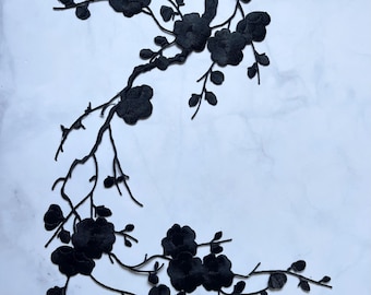 May include: Black floral embroidery applique featuring a branch with flowers and leaves. The applique is made of a textured fabric and is set against a white marble background. The design is detailed and intricate, suitable for embellishing garments or crafts.