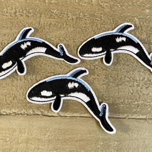 May include: Three black and white embroidered iron-on patches of orca whales. The whales are facing different directions.