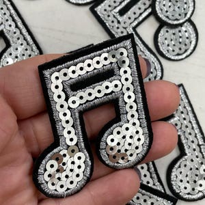 Op de afbeelding: Zilveren pailletten muzieknoot patches op een zwarte stoffen achtergrond. De patches zijn ongeveer 5 cm groot.