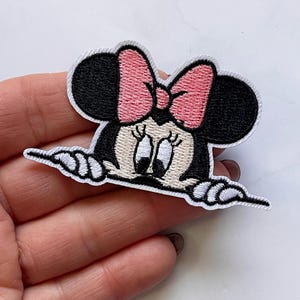 Könnte beinhalten: Gestickter Bügel-Patch mit einem Cartoon-Bild von Minnie Mouse, die über eine weiße Oberfläche schaut. Minnie Mouse hat eine rosa Schleife und schwarze Ohren.
