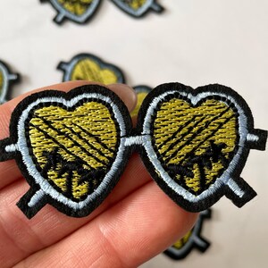May include: A pair of heart-shaped sunglasses with a yellow and black palm tree design. The sunglasses are embroidered with black thread and have a light blue outline.