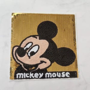 May include: Square gold sequin patch featuring Mickey Mouse. The cartoon character is black, white, and pink. The words "mickey mouse" are below the character in white letters.