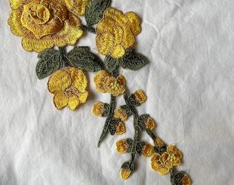 May include: Embroidered floral applique featuring yellow roses and green leaves. The detailed design showcases a cluster of flowers with intricate stitching, perfect for embellishing clothing or crafts. The applique is set against a white background.