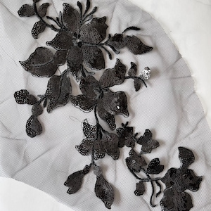 May include: Black sequined floral applique on a gray mesh fabric. The applique features detailed flower and leaf designs, perfect for embellishing garments or accessories. The sequins add a shimmering effect to the floral pattern.