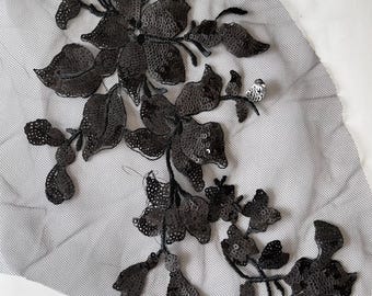 May include: Black sequined floral applique on a gray mesh fabric. The applique features detailed flower and leaf designs, perfect for embellishing garments or accessories. The sequins add a shimmering effect to the floral pattern.