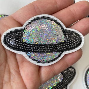 Puede incluir: Un parche de planchado con lentejuelas plateadas y negras en forma de planeta con anillo. El parche está rodeado por un borde blanco.