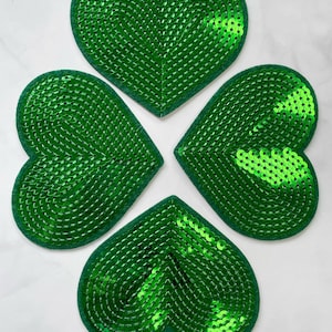 Puede incluir: Cuatro parches verdes con forma de corazón y lentejuelas, dispuestos como un trébol de cuatro hojas. Las lentejuelas verdes vibrantes destacan sobre un fondo blanco. El trébol es un símbolo de buena suerte.
