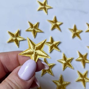 May include: Close-up of several gold star-shaped embroidered patches. The patches are scattered on a white surface. One star is held by a person. The stars have a shiny, metallic appearance.