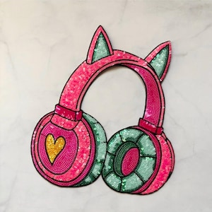 May include: A pink sequined headphone patch with cat ears. The ear cups are embellished with green sequins and a gold heart. The headphones are outlined in black and set against a white background.