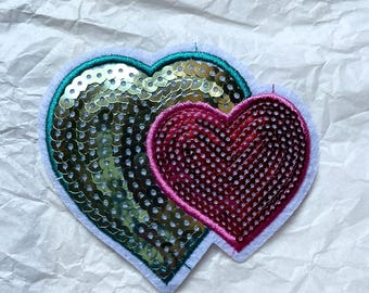 Puede incluir: Dos parches en forma de corazón. Un corazón es dorado con ribete turquesa y el otro es rojo con ribete rosa. Ambos corazones están cubiertos de lentejuelas y sobre un fondo blanco.