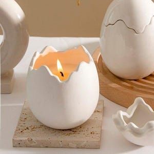 May include: A white ceramic egg-shaped candle holder with a lit candle inside, sitting on a square stone coaster. Another egg-shaped container with a lid is on a wooden tray. A small, white, flower-shaped dish is also present.