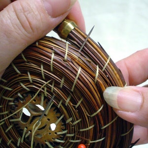 May include: Close-up of a woven basket with a brown and tan pattern. The basket is being held by a hand and has a brass pin inserted into the top.