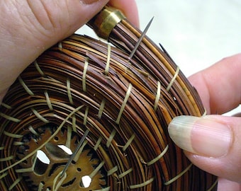 May include: Close-up of a woven basket with a brown and tan pattern. The basket is being held by a hand and has a brass pin inserted into the top.