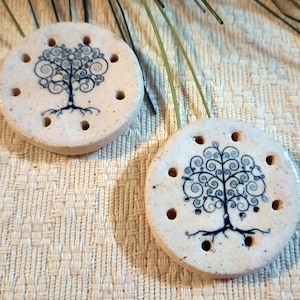 May include: Two round, off-white ceramic pieces with a blue tree design. Each piece has eight small holes around the edge. The tree design features a detailed trunk, branches, and leaves.